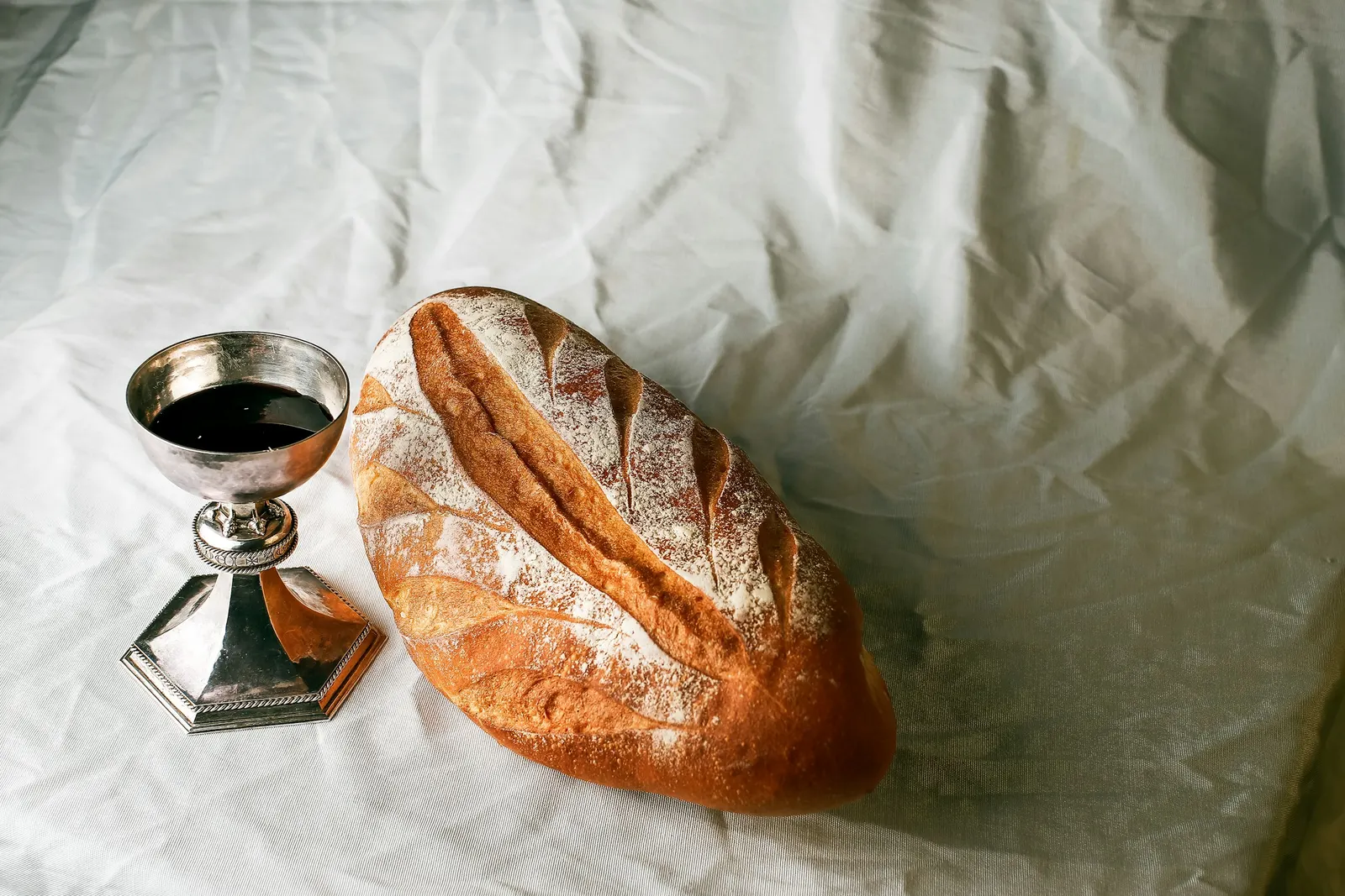 a loaf of bread beside a polished silver chalice of wine on white linen