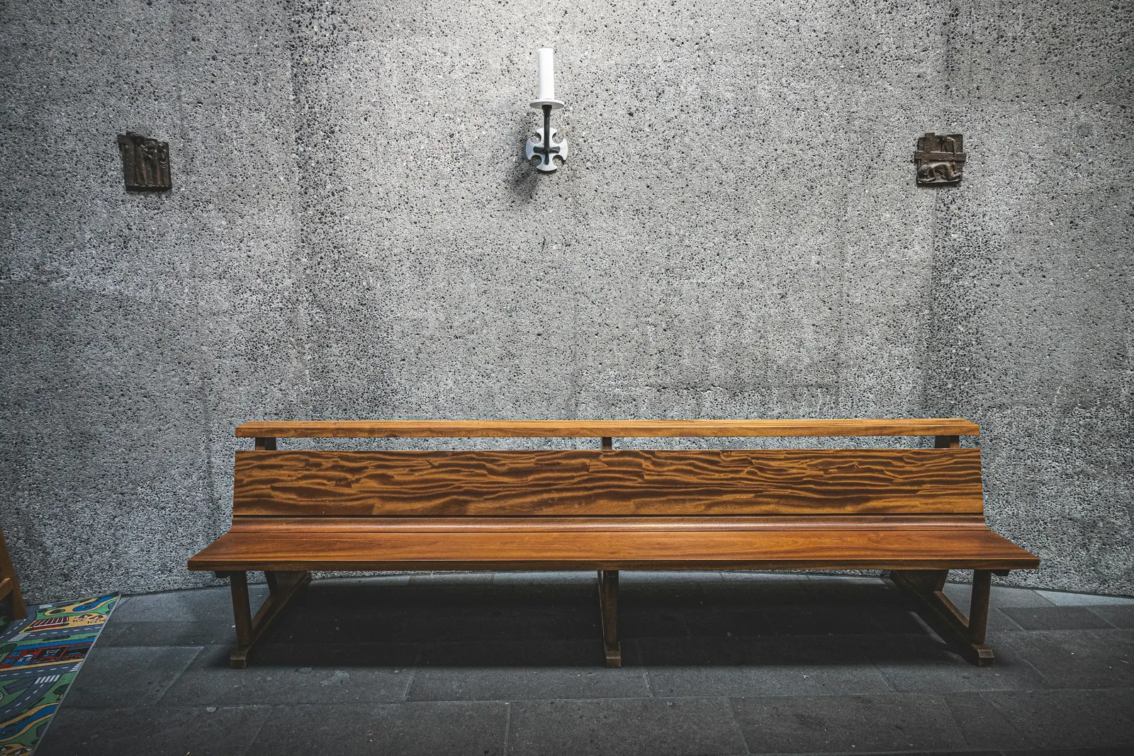 a smooth wooden pew bench against a bare concrete wall with a small candle sconce above it